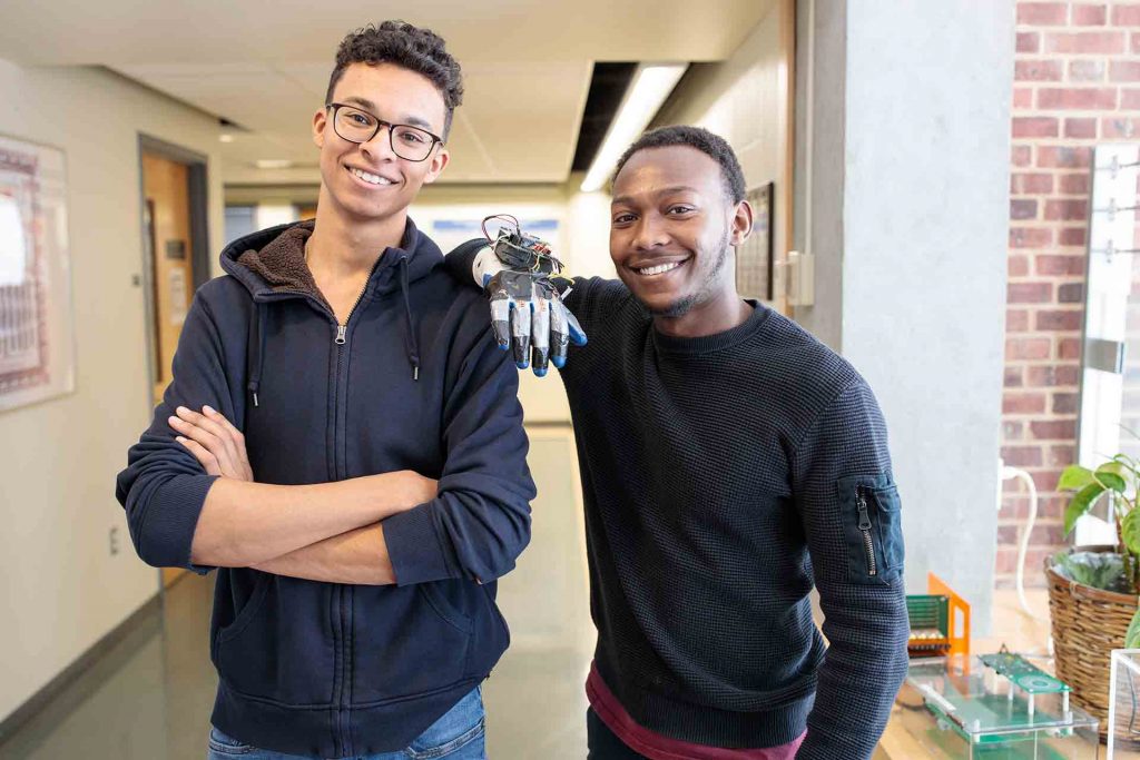 Two male students pose with their glove. One leans on the other who has his arms crossed.