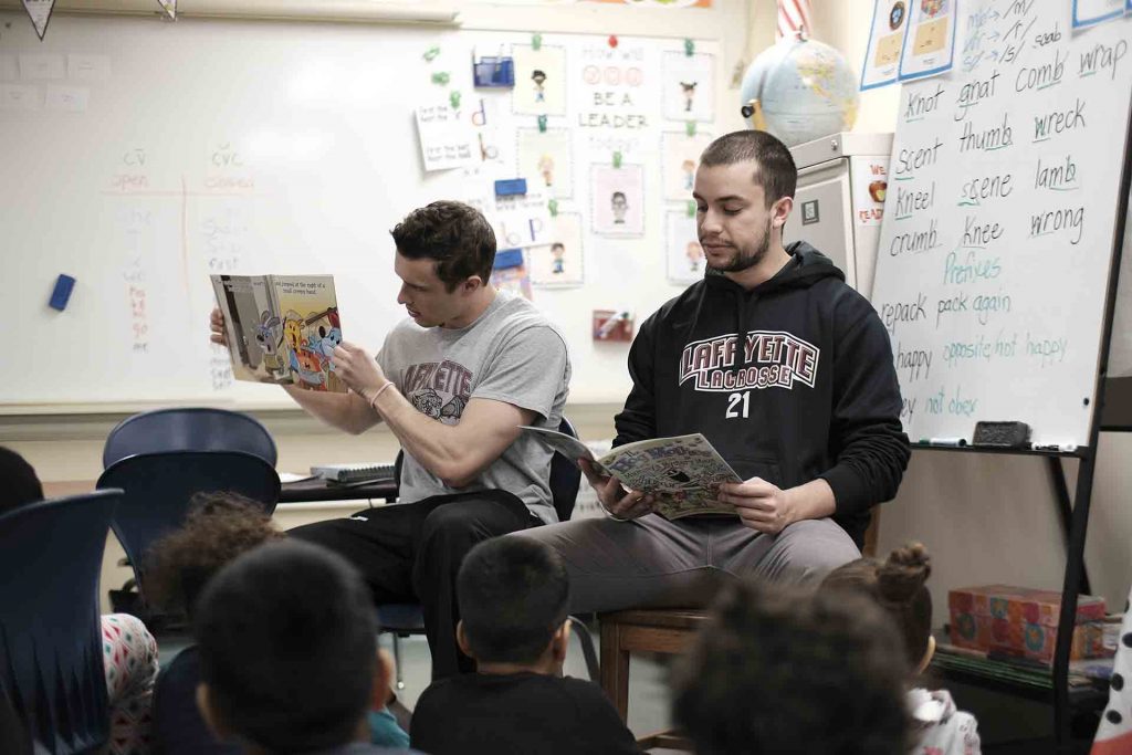Two lacrosse players take turn reading a book to second graders.