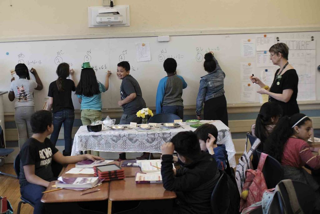 5th grade students stand at a whiteboard and write their math problems. One boy is turned around and making a funny face.