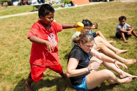 A kid in the community squeezes a wet sponge on a college student's head