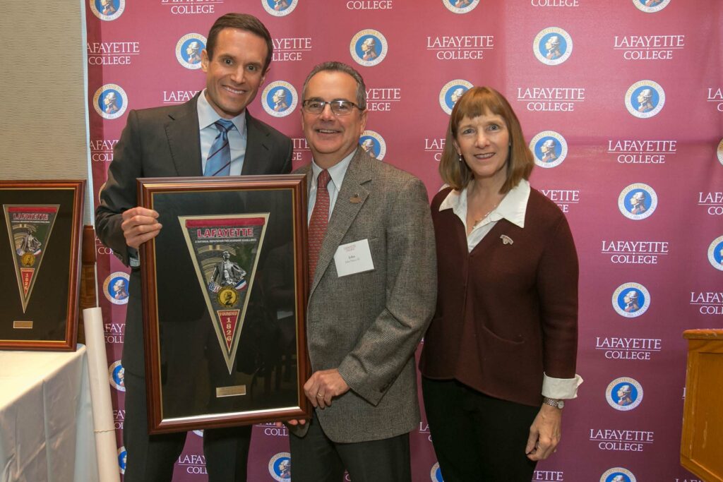 John holds a framed award and is flanked by the alumni and college presidents