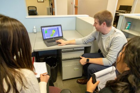 Student externs sit at desk with host who shows them something on a computer