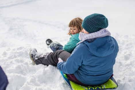 an adult and child sit on a sled in the snow on campus