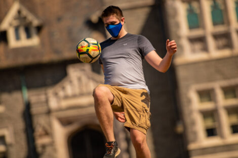 A masked student kicks a soccer ball with Hogg Hall behind them.