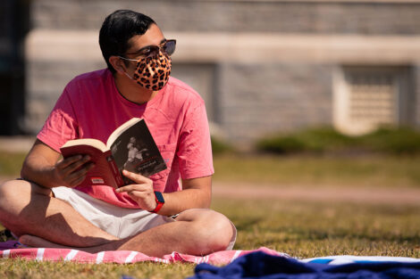 A masked student holds open a book, sitting on a blanket on the grass of the Quad.
