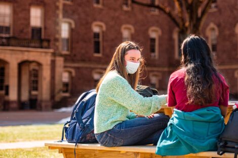 A masked student talks to another masked student, sitting on a bench outside of Pardee Hall.