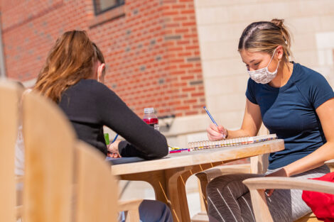 Two masked students sit opposit of each other at a table outside of a brick building.