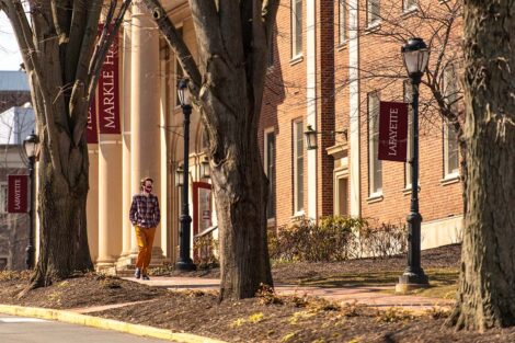 A masked student walks outside of Markle Hall. Trees, lamp post, and Lafayette flags line the sidewalk
