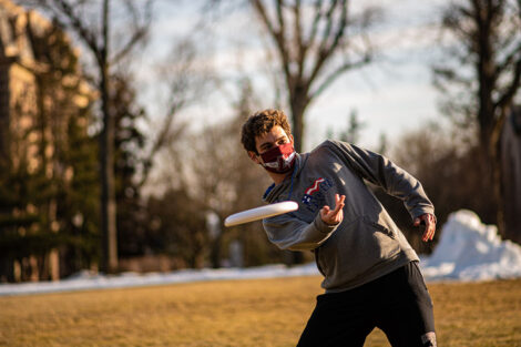 A masked student throws a frisbee on the Quad outside of Pardee Hall