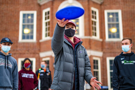 A student wearing a mask tosses a frisbee with other students standing behind them.