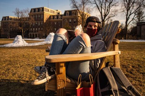 A masked student sits in an Adirondack chair with knees up, on the Quad in front of Pardee Hall.