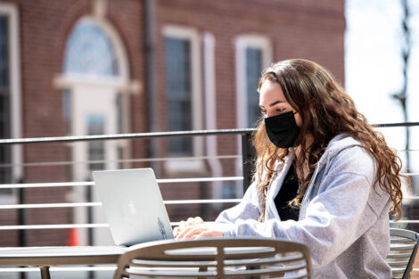 A masked student sits outside of Rockwell Integrated Sciences Center, with their laptop open.