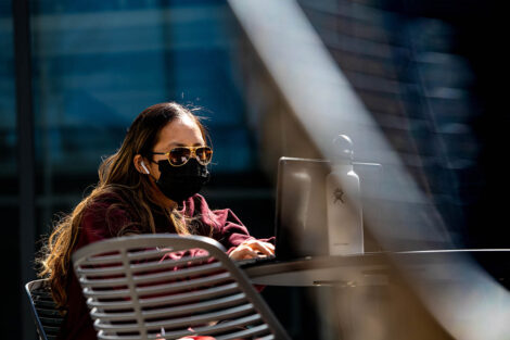 A masked student sits at a table outside, typing on their laptop; a reusable water bottle is also on the table.