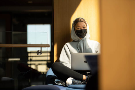 A masked student, with their sweatshirt hood up, sits with their laptop open inside of Rockwell Integrated Sciences Center