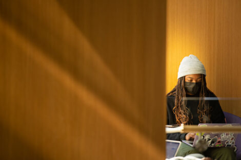 A masked student in a hat, sits in front of their laptop in Rockwell Integrated Sciences Center
