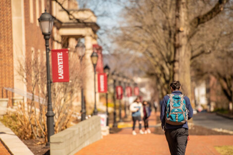 Students walk away from the camera on a brick sidewalk, lined with lamp posts and Lafayette College flags, in front of Markle Hall.
