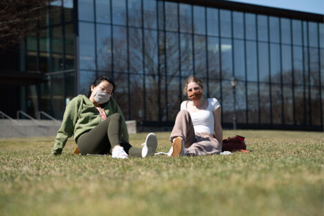Two masked students sit in grass in front of Skillman Library.