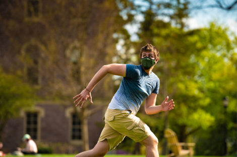 A masked student runs to retrieve a frisbee.