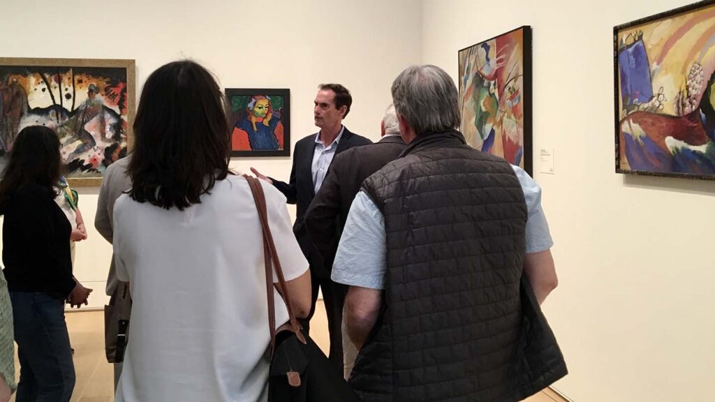 Bob Mattison speaks to alumni in a gallery with several paintings on the wall behind him