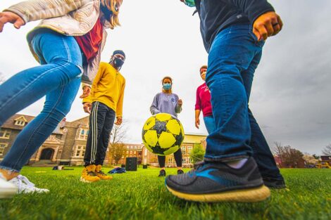 Five masked students stand in a circle, kicking a ball in front of Hogg Hall and Farinon College Center