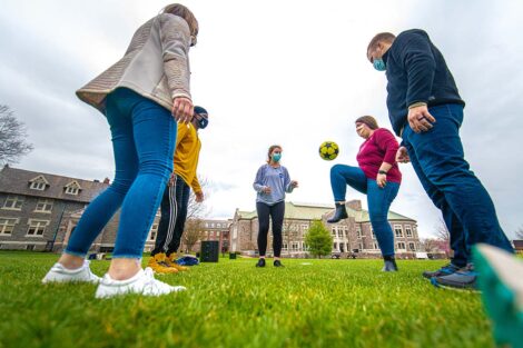 Five masked students stand in a circle, kicking a ball in front of Hogg Hall and Farinon College Center