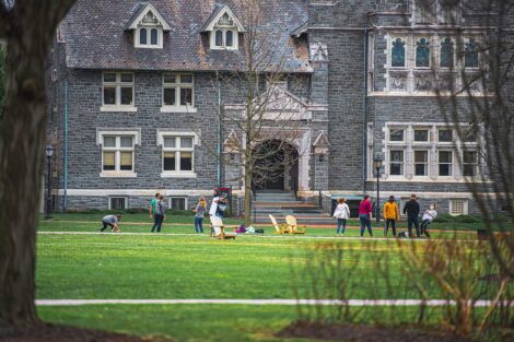 Five masked students stand in a circle, kicking a ball in front of Hogg Hall and Farinon College Center