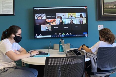 Two students sit at a table. A wall-mounted TV displays their laptop screens and shows a group of participants on Zoom for Literacy Day