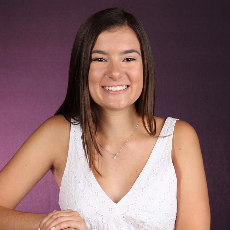 Maura DiGuglielmo stands before a purple backdrop and wear a white sundress