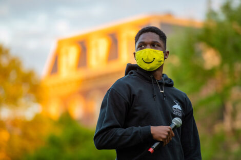 A student, wearing a yellow smiley face mask, holds a microphone in front of Pardee Hall.