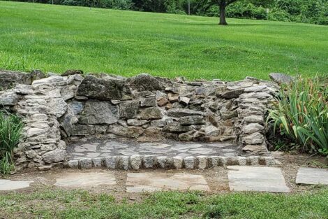 A reconstructed firepit with three sides of rock wall sits in the side of a green hill