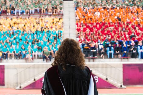 President Hurd looks out at the Fisher Field stadium seats, filled with students, students sit in groups with yellow, red, blue, and orange Tshirts