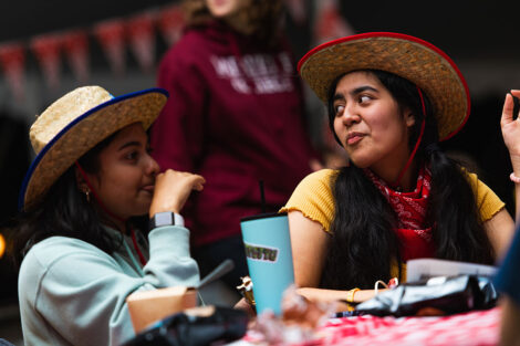 Two students in cowboy hats and bandanas sit at a covered picnic table.