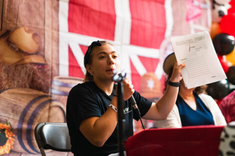 A student speaks into a microphone and holds a trivia sheet underneath a tent in the Quad.