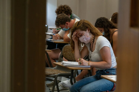 A student, wearing a mask, takes notes in a classroom, surrounded by other students.