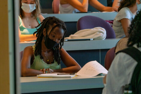 A student, wearing a mask, looks over notes at a desk inside of a classroom.