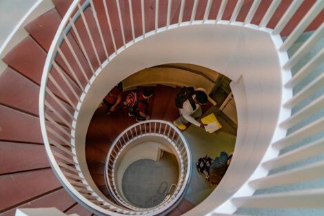 A spiraling staircase. Students, wearing masks and backpacks and carrying books, walk up them.