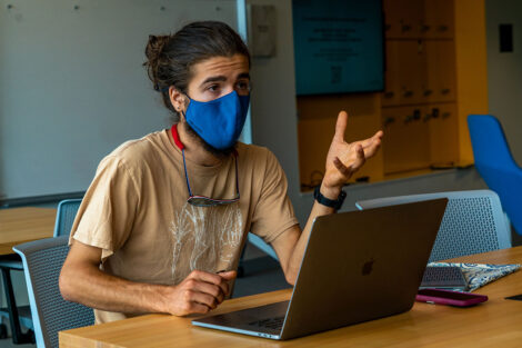 A student, wearing a mask, engages in conversation with their laptop open.