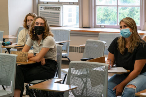 Students, wearing masks, sit in desks inside of a classroom.
