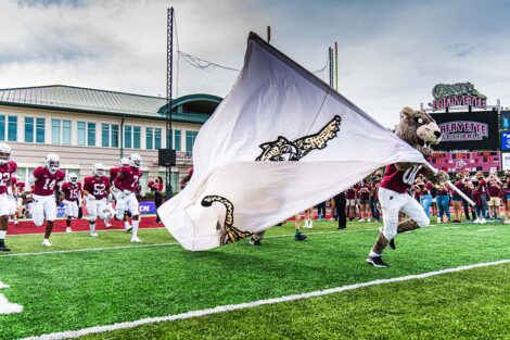 The Lafayette Leopard runs across Fisher Field, carrying a flag with a Leopard on it. Football players run behind.