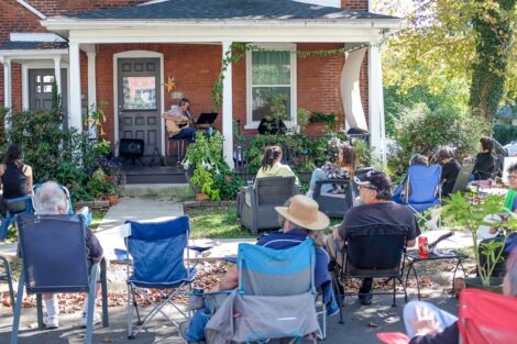 performer sings and plays guitar on a porch with audience in folding chairs