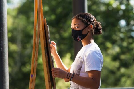 Students stands at easel with greenery behind her
