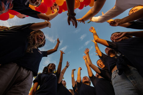 students gather in a circle and raise hands toward blue sky
