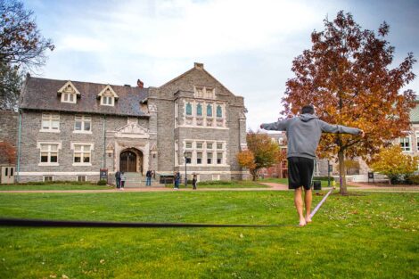 student balances on a tight robe attached between two trees on the Quad, Hogg Hall in background