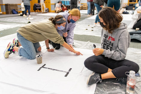 Students, wearing masks, paint on white banners.