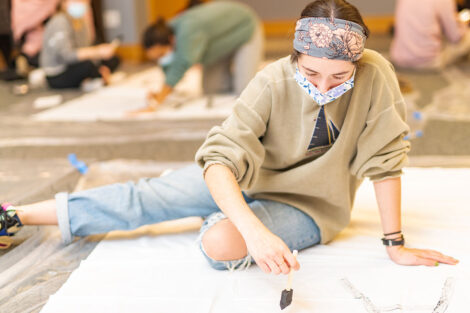 Students, wearing masks, paint on white banners.