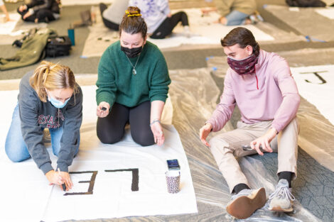 Students, wearing masks, paint on white banners.