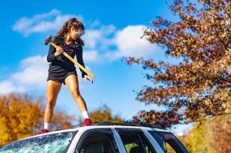 A student stands on top of an SUV and hits it with a sledgehammer.