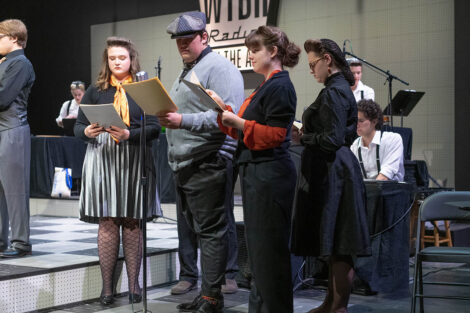 student performers in vintage dress gather on stage with vintage microphones