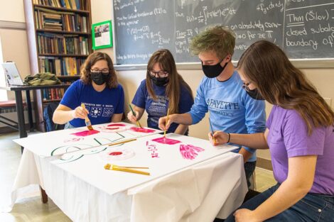 group of students sit on floor and paint sign, blackboard filled with math equations is behind them