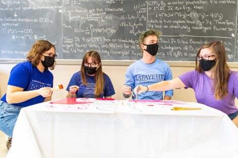 group of students sit on floor and paint sign, blackboard filled with math equations is behind them
